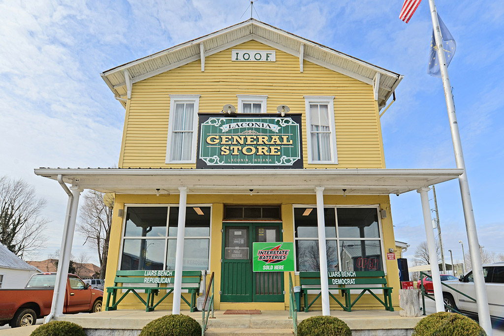 Close up the restored 1900s Laconia General Store. Soft yellow exterior with white trimmed windows, green doors and benches, and circular cut hedges in the front landscaping.